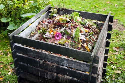 A black plastic compost bin situated on a grassy area with small patches of dirt, filled with organic waste including plant matter, leaves, and flowers, which appear damp and decomposing. Surrounding the bin are green leafy plants, and the background features a lawn with scattered foliage. The scene is outdoors, suggesting a garden or backyard environment, with natural daylight illuminating the waste and foliage. This image illustrates waste suitable for collection and removal by a rubbish clearance service like Commercial Waste Leyton, serving Leyton and nearby postcode areas, with the compost bin reflecting environmentally focused waste management practices in London.