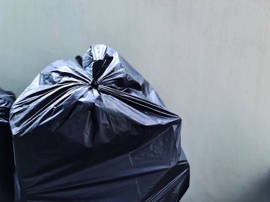 A pile of discarded rubbish situated on a gravel surface outdoors, consisting of mixed waste including black and white plastic rubbish bags, some filled with unknown refuse, and a yellow plastic conta
