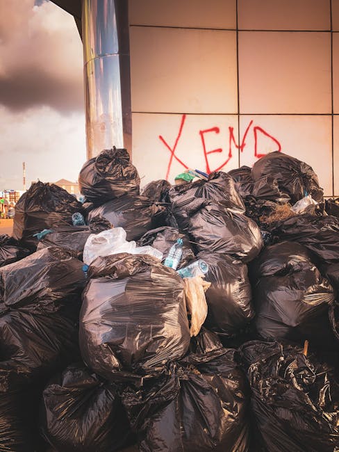 A large pile of black plastic rubbish bags, some with visible creases and twists, stacked against a tiled wall. The bags appear full and bulky, with a few partially torn or crumpled, revealing occasio